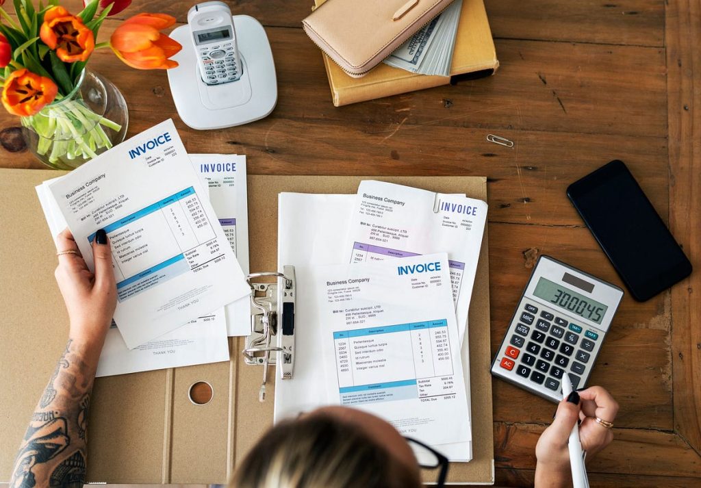A person reviewing invoices and using a calculator while surrounded by office supplies, including a phone and a flower vase.