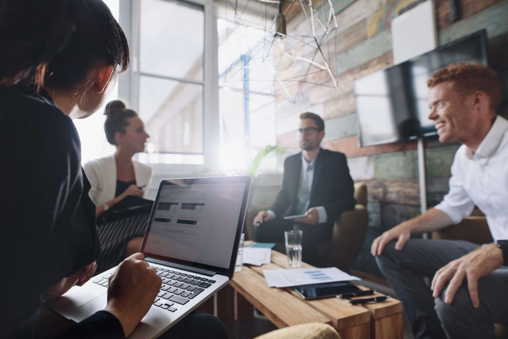 A group of professionals in a meeting with one person working on a laptop and others engaged in conversation.