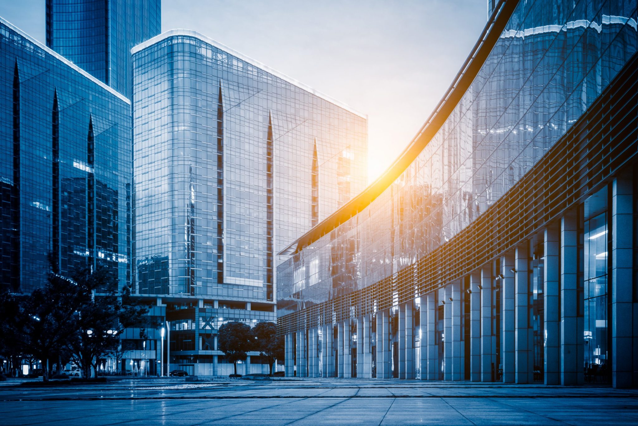 Modern glass office buildings and plaza at sunset with warm sunlight creating golden reflections on facades.