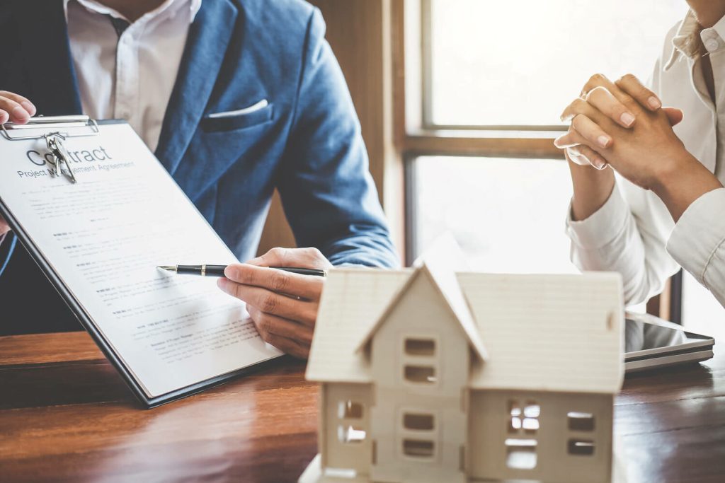 A person pointing at a contract while another person sits with their hands clasped, with a house model on the table.