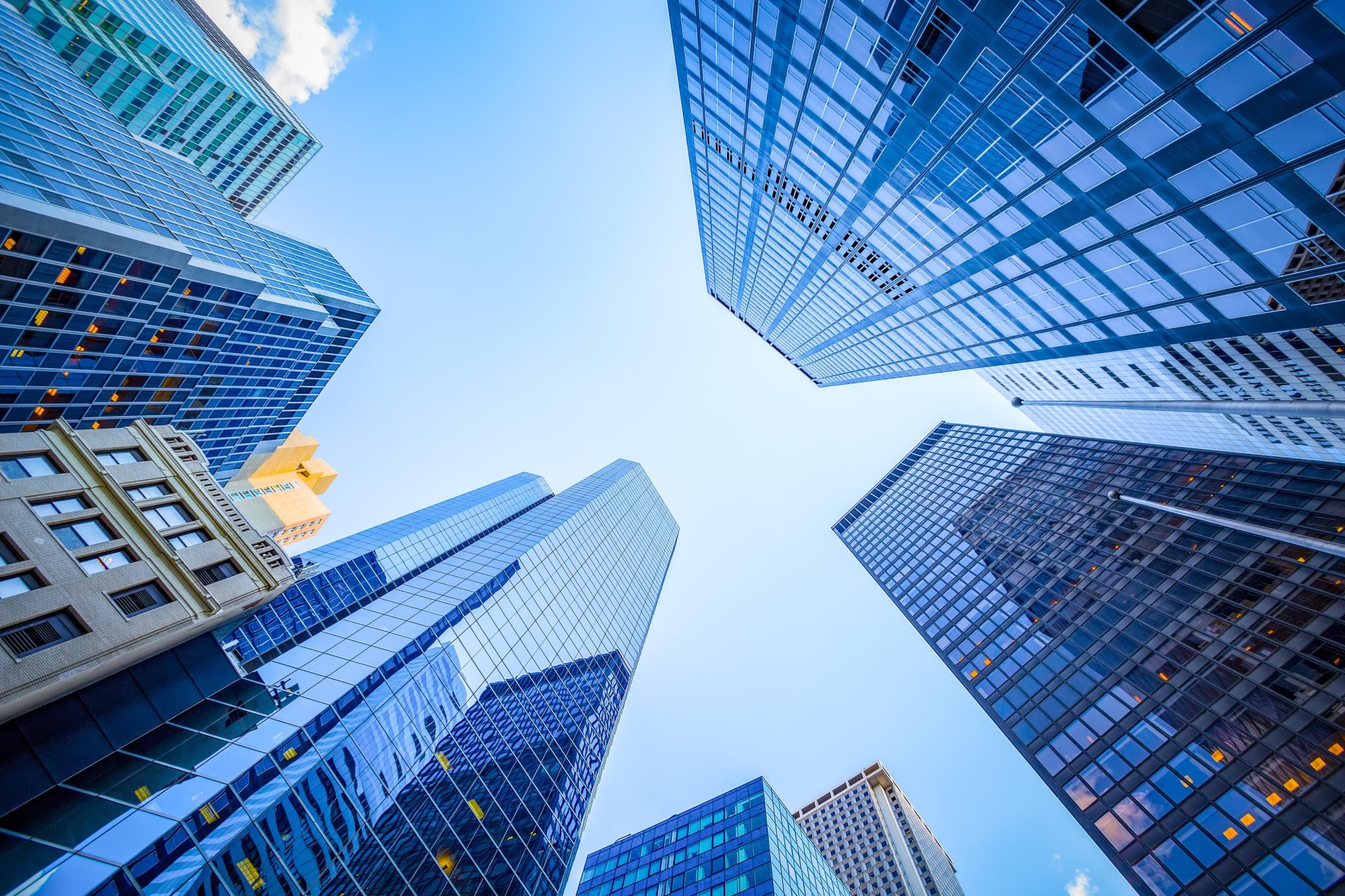 Upward view of modern glass office towers with blue sky and clouds, showing urban financial district architecture.