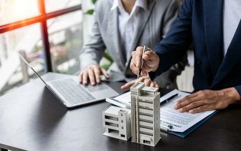 Two people in business attire discussing a building model, with one signing a document and the other using a laptop.
